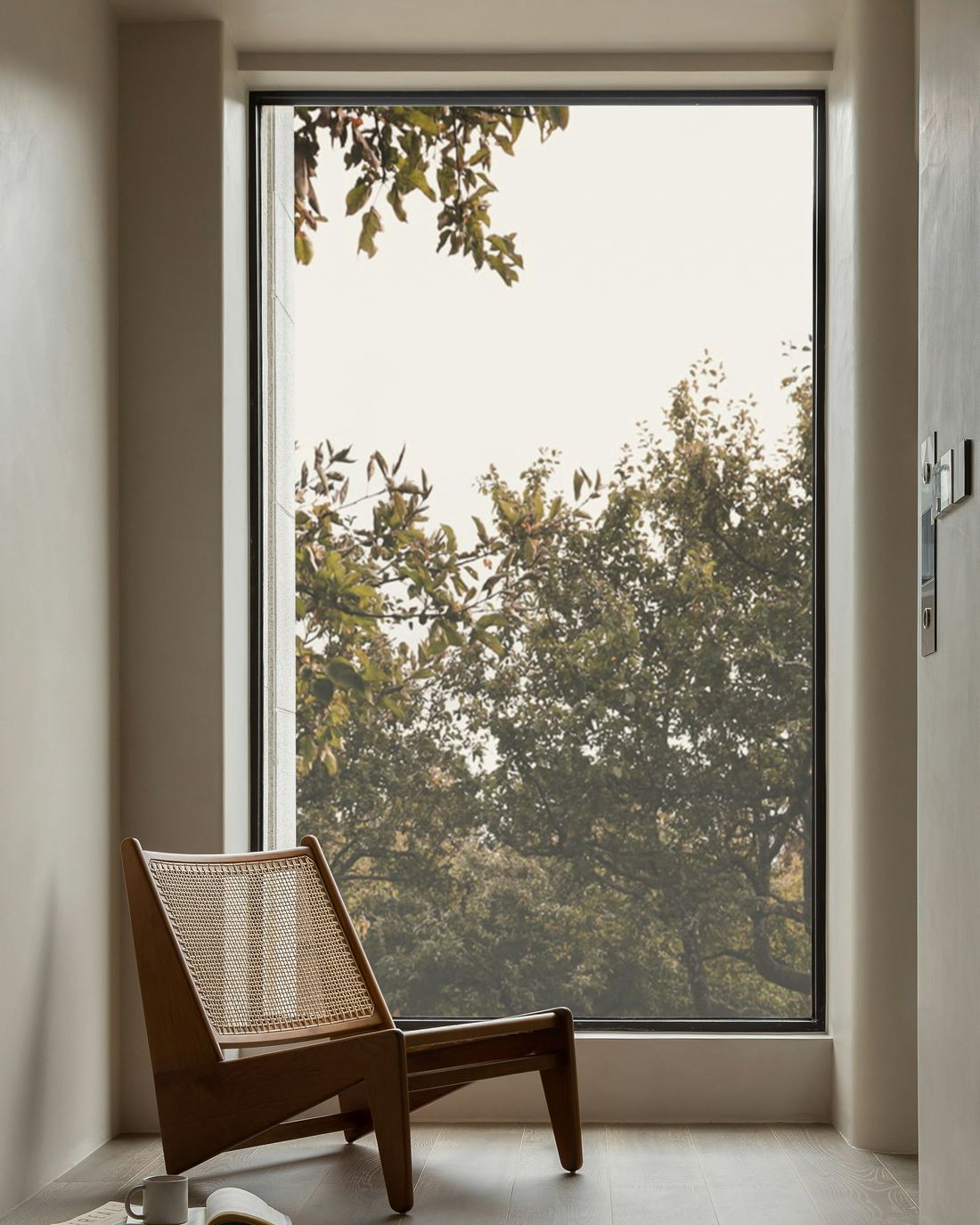 A cane chair by a tall window with books and a mug on the floor